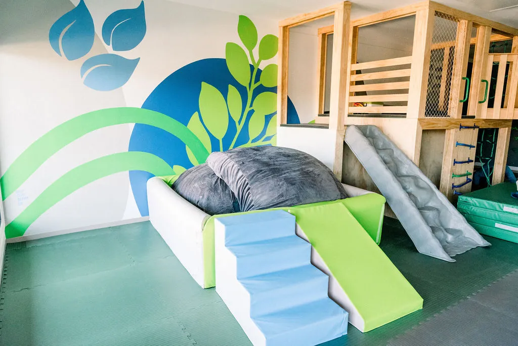 Soft play area with gray cushions, blue steps, and a lime green slide beside a wooden climbing structure