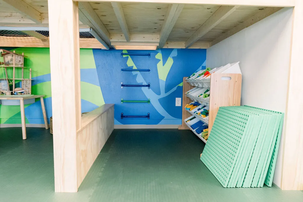 Play nook under a wooden loft with a toy organizer, green mats, and blue climbing rungs