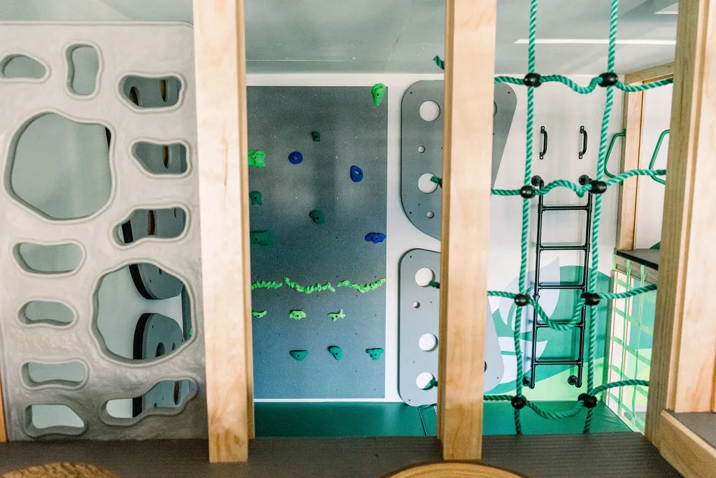 Indoor climbing wall with grey bouldering holds, a green rope cargo net, and a black metal ladder