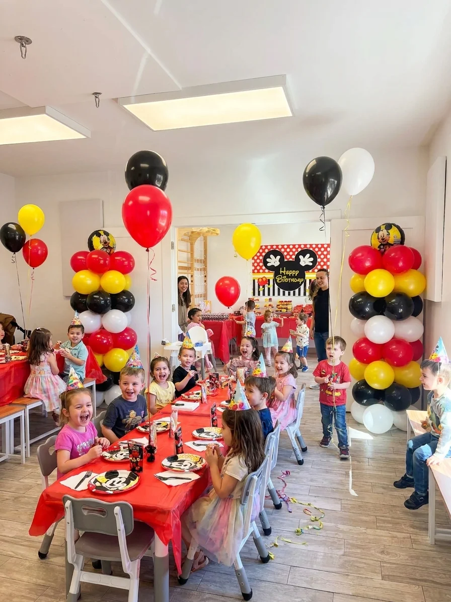 Children in party hats at a Mickey Mouse-themed table with red, yellow, and black balloon pillars